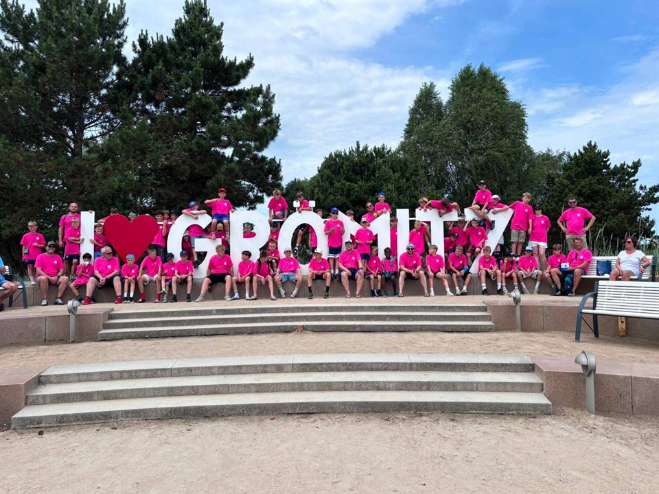 Große Gruppe in pinken Vereinsshirts vor dem Schriftzug „I love Grömitz“ am Strand