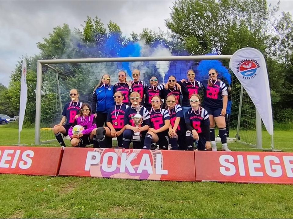 Mädchenmannschaft in dunkelblauen Trikots mit pinken Nummern, Medaillen und Pokal vor dem Tor am Banner „Kreispokalsieger“